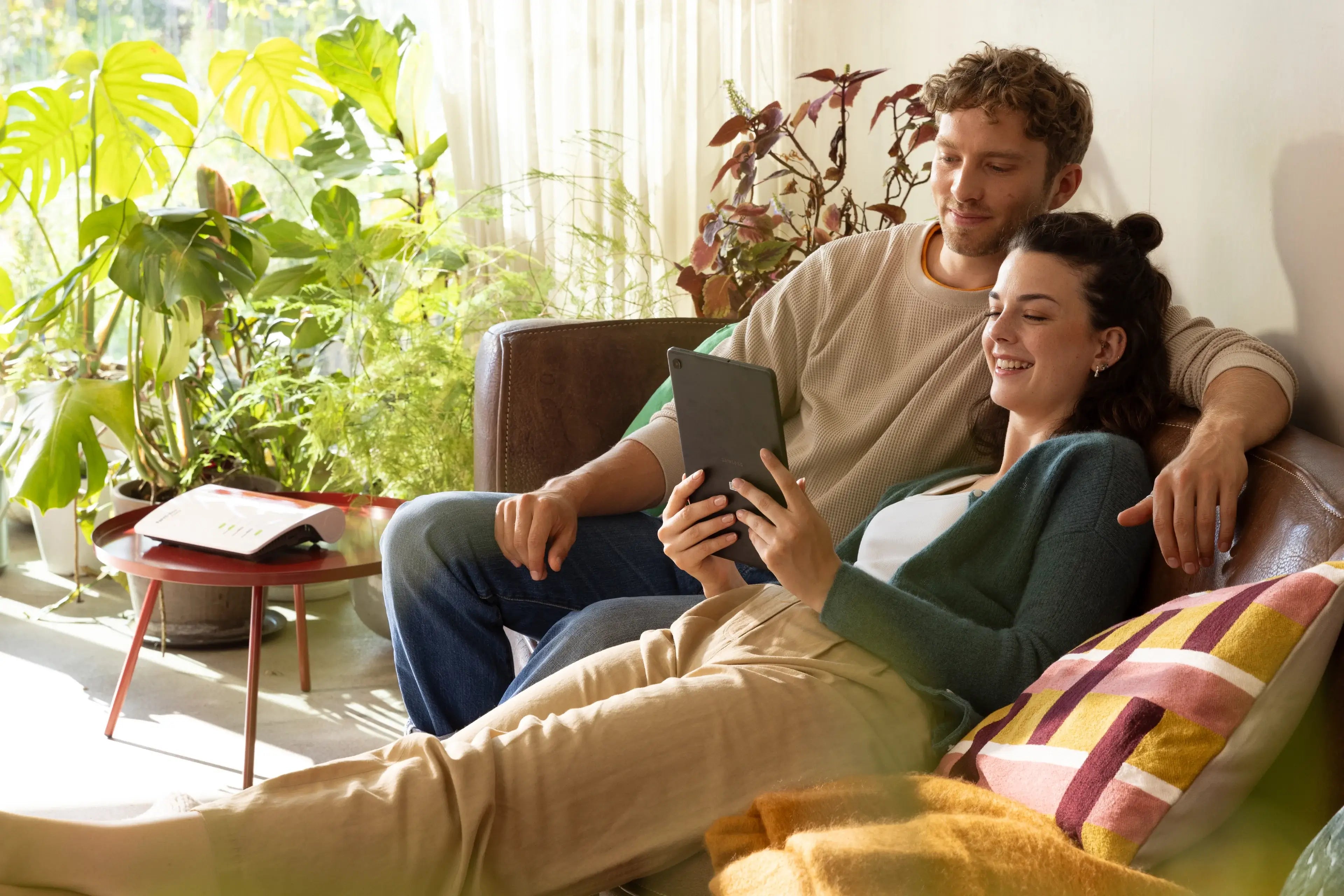 Ein Paar entspannt auf einer Couch und schaut auf ein Tablet, umgeben von Pflanzen und gemütlicher Einrichtung.