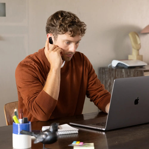 Mann mit Locken nutzt kabellose Ohrhörer und arbeitet am Laptop in einem modernen Büro.